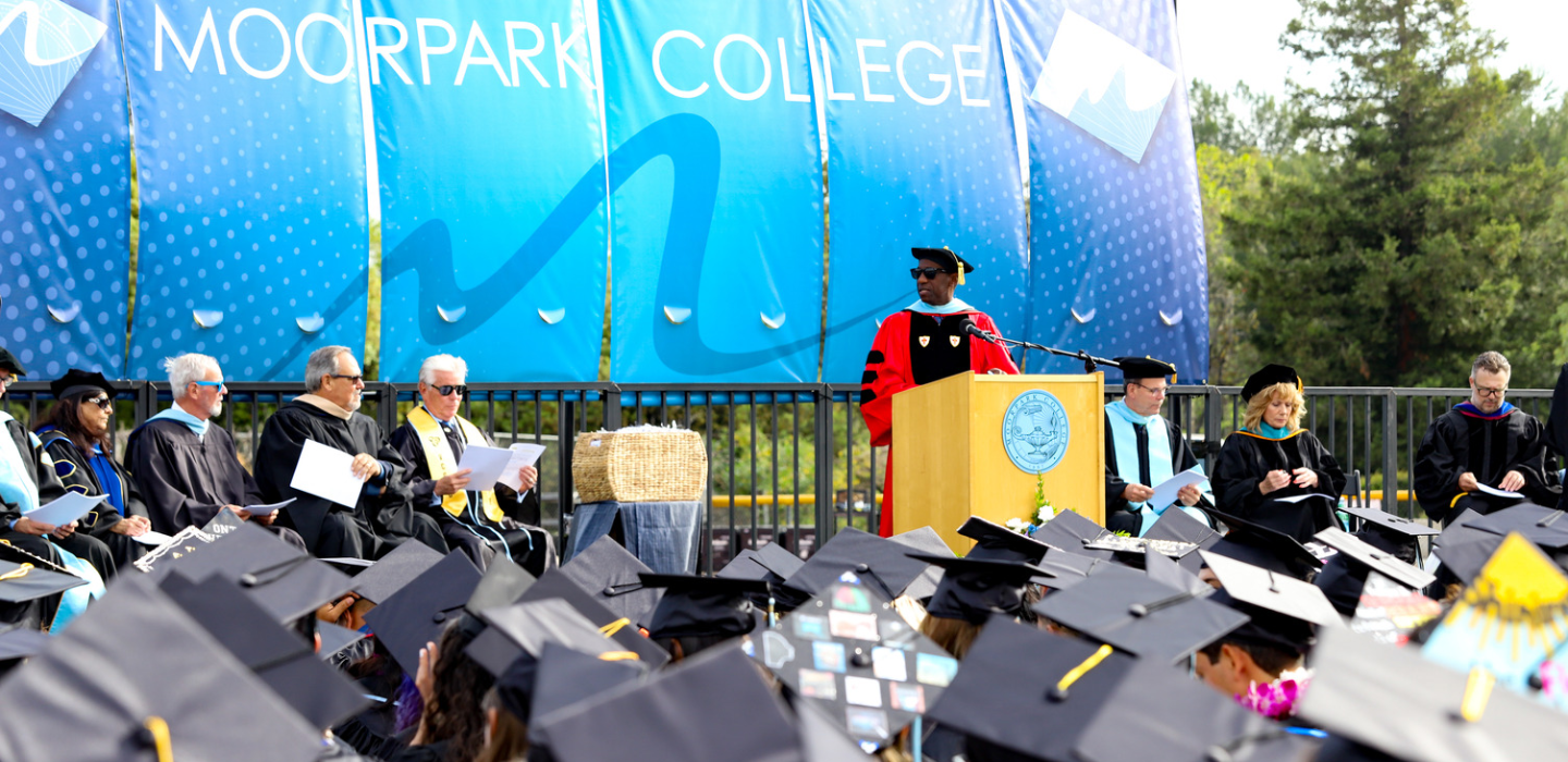 Dr. Sokenu stands in front of lectern on stage with Moorpark College written on the banner behind him facing out towards the graduates.