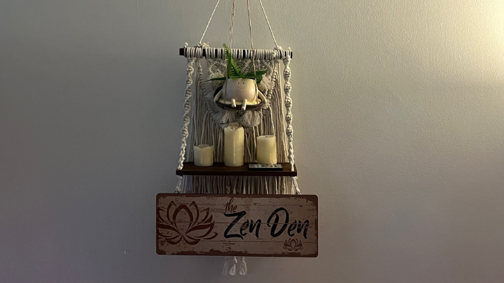 Macrame shelf with candles is shown with a sign that reads the Zen Den hanging from it.